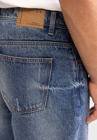 Jean en denim de couleur bleu clair délavé, avec des coutures orange et une patte en cuir marron au niveau de la taille, comportant un texte.