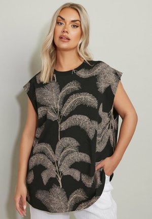 Donna con capelli biondi che indossa una canotta nera con stampa di foglie di palma color sabbia e pantaloni bianchi, posando con una mano in tasca contro un muro semplice.