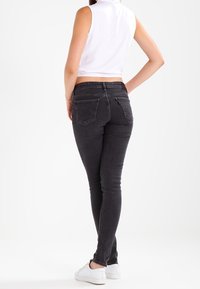 Jeans skinny negros con cintura de altura media, bolsillos traseros y detalles cosidos, combinados con una camiseta blanca sin mangas y recortada, y zapatillas blancas.