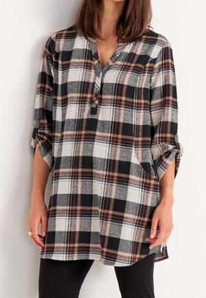 Kobieta ubrana w luźną, czarno-biało-brązową koszulę w kratę z długim rękawem, częściowo zapinaną z przodu na guziki i podwiniętymi rękawami.