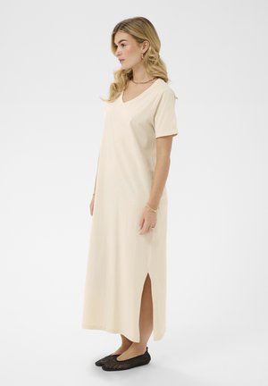 Pikkade blondide juustega naine kreemika värvi V-kaelusega poolpikk kleit, millel on lühikesed varrukad ja mustad kootud madalad kingad, seisab külje pealt.