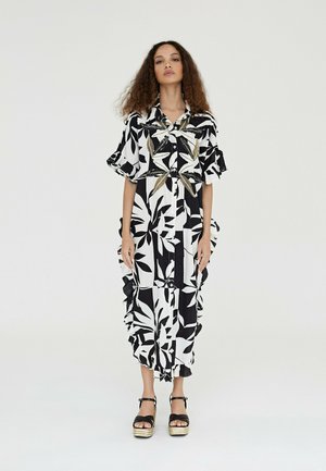 Vestido floral en blanco y negro con mangas cortas con volantes, botón frontal y dobladillo con volantes. Llevado con sandalias de plataforma negras y acentos dorados.