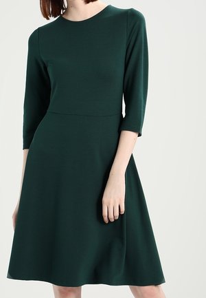Kvinde iført en mørkegrøn kjole til knæet med trekvartærmer og en tilpasset talje, stående mod en ensfarvet lys baggrund.