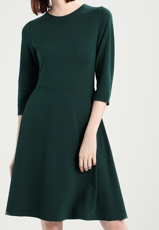 Femme portant une robe vert foncé longueur genou avec des manches trois-quarts et une taille cintrée, debout devant un fond clair uni.