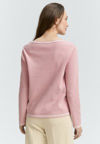 Maglione in maglia rosa chiaro con righe orizzontali, maniche lunghe e scollo ampio. Texture liscia e polsini a coste, indossato sopra pantaloni beige.
