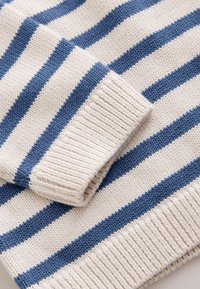 Manche de pull tricoté à rayures crème et bleu repliée sur le corps du pull avec des poignets et un bas côtelés.