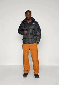 Schwarze Pufferjacke mit Reißverschluss und Kapuze, kombiniert mit orangen Hosen. Verfügt über sichtbare Nähte und strukturierte Materialien. Schwarze Stiefel ergänzen das Outfit.