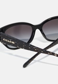 Schwarze Cat-Eye-Sonnenbrille mit verlaufenden Gläsern. Acetatrahmen mit einem Schildpatt-Muster an den Bügeln. Goldenes "COACH"-Logodetail.