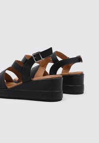 Sandalias de cuña de cuero negro con una suela estratificada, correas ajustables con hebilla y un forro interior suave en tono marrón.