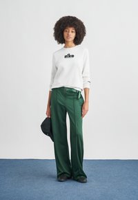 Γυναίκα με σγουρά μαλλιά που φορά λευκό φούτερ Nike, πράσινο παντελόνι με φαρδιά μπατζάκια, μαύρα παπούτσια, κρατώντας μαύρο καπέλο δίπλα σε λευκό τοίχο και μπλε πάτωμα.