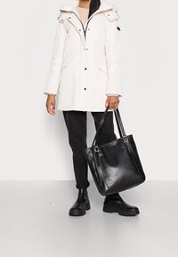 Puchowa biała parka z kapturem, czarnymi guzikami, kieszeniami bocznymi, w zestawieniu z czarnymi spodniami i dużą czarną skórzaną torbą. Grube czarne botki.