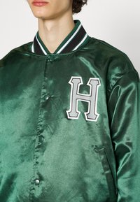 Grüne Satin-Baseballjacke mit schwarzem und weißem gestreiftem Rippkragen. Sie verfügt über einen auffälligen grauen Buchstaben "H" und Knopfverschlüsse auf der Vorderseite.