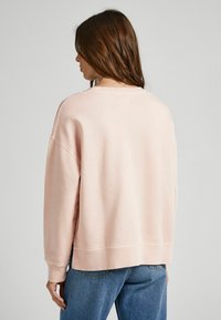 Felpa rosa chiaro con vestibilità ampia, spalle basse e collo a giro. Texture morbida, polsini e orlo a costine. Abbinata a jeans blu.