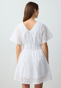 Weißes Baumwollkleid mit V-Ausschnitt, kurzen Ärmeln, elastischem Bund und kreisförmigen Lochmustern im gesamten Stoff.