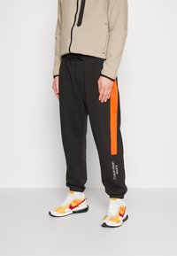 Black sweatpants feature an orange stripe along the side and “Calvin Klein Jeans” logo. Paired with white and yellow athletic shoes.
