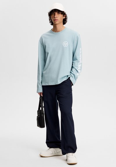 Lichtblauw overhemd met lange mouwen en een logo op de borst, gecombineerd met marineblauwe broek. Model draagt een zwarte tas, witte sneakers en een witte pet.