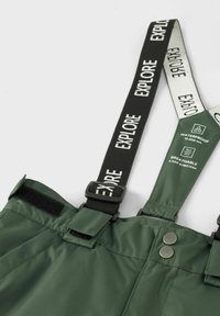 Vandeniui atsparūs žali audiniai overallai su reguliuojamomis juodomis petnešomis, turinčiais tekstą „TIRKTI“, Velcro užsegimus ir metalinius sagos akcentus.