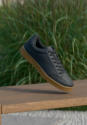 Sort lædersneaker med gummisål, der svæver over en træbænk med høj grønt græs i baggrunden.