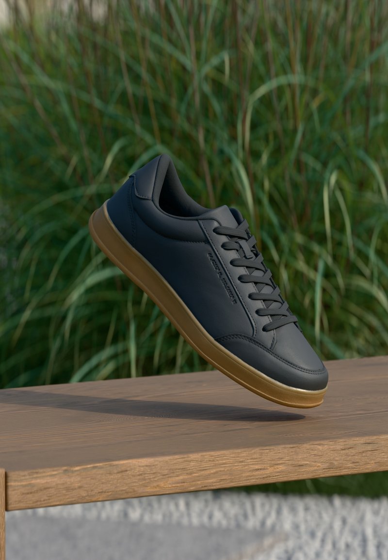 Baskets en cuir noir avec semelle en gomme flottant au-dessus d'un banc en bois, avec de hautes herbes vertes en arrière-plan.
