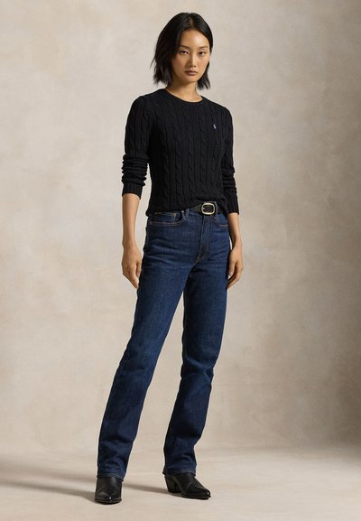 Nainen seisoo päällään musta palmikkoneulepaita, tummansiniset farkut vyöllä ja mustat korkokengät neutraalia taustaa vasten.