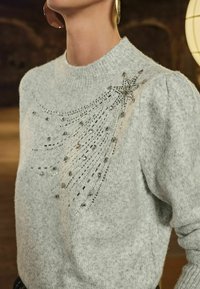 Pilkas megztinis su raukšlėtu kaklu, dekoruotas žvaigždžių rašto karoliukais, pagamintas iš minkštos, tekstūruotos medžiagos.