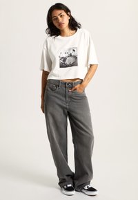 Witte cropped t-shirt met een zwart grafisch ontwerp. Gecombineerd met hoge, losse grijze jeans en zwart-witte sneakers.