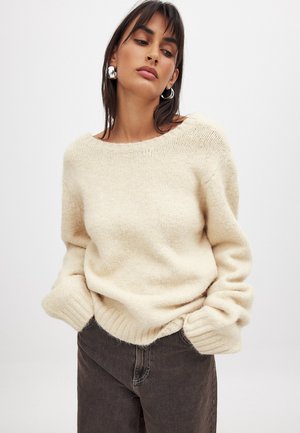 Beige, gebreide trui met een wijde halslijn en geribbelde manchetten, gecombineerd met donkerbruine broek. Zachte, gestructureerde stof zichtbaar.