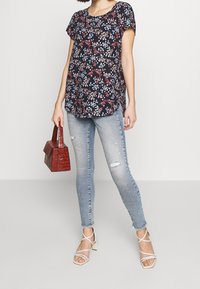 Námořnické květinové tričko s krátkými rukávy a kulatým výstřihem, v kombinaci se světle modrými skinny džínami, červenou texturovanou kabelkou a sandály na podpatku v neutrální barvě.