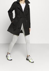 Persona che indossa un cappotto nero con cintura, una camicia bianca, leggings grigio chiaro e sneakers bianche con dettagli neri e neon verde.