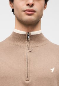 Pull beige à fermeture éclair avec texture côtelée, col montant et accent crème. Comprend une fermeture éclair contrastante et un petit logo d'oiseau blanc.