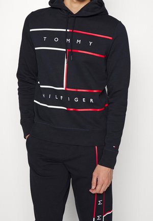Homme portant un sweat à capuche noir Tommy Hilfiger et un pantalon assorti avec des bandes blanches et rouges et un lettrage de logo.