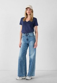 Mujer con una gorra beige, camiseta de cuello en V azul marino, vaqueros de talle alto y pernera ancha azules, y zapatillas plateadas, de pie frente a una pared lisa.