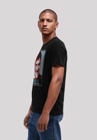 Man with shaved head wearing black graphic t-shirt and blue jeans, standing sideways against a plain light gray background.