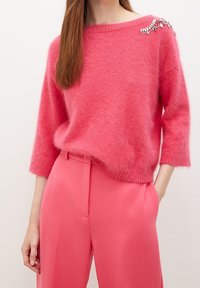 Pehmeä vaaleanpunainen neule leveällä pääntiellä ja kolme neljäsosaa hihoilla, koristeltu vasemmalla olkapäällä jalokivikoristein, yhdistetty vaaleanpunaisiin housuihin.