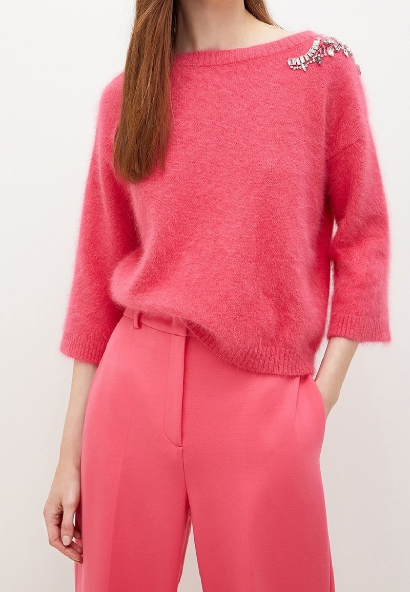 Pehmeä vaaleanpunainen neule leveällä pääntiellä ja kolme neljäsosaa hihoilla, koristeltu vasemmalla olkapäällä jalokivikoristein, yhdistetty vaaleanpunaisiin housuihin.