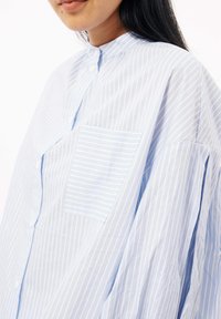 Lichtblauw gestreept oversized shirt met een bandkraag, sluiting met knopen aan de voorkant en een enkele borstzak met horizontale strepen.