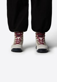 Bottes de randonnée gris clair avec des lacets bordeaux et des œillets dorés, dotées d'une semelle extérieure noire robuste, conçues pour les activités en plein air.