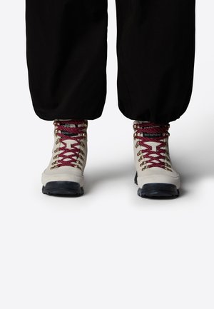Bottes de randonnée beiges avec des lacets rouges, des semelles noires et des œillets métalliques, portées avec un pantalon noir sur fond blanc.