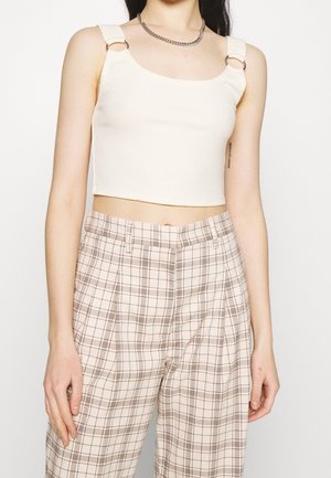 Mujer que lleva un top corto de canalé color crema con detalles de anillos en las tiras y pantalones beige a cuadros de talle alto sobre un fondo liso.