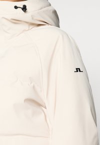 Ανοιχτό καφέ μαλακή ζακέτα διαθέτει ψηλό γιακά, ρυθμιζόμενα κορδόνια και μαύρη λογότυπο ένδειξη στο μανίκι.