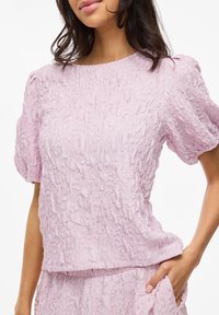 Top rosa chiaro testurizzato con maniche a sbuffo corte e scollo rotondo, caratterizzato da un motivo di tessuto stropicciato e una vestibilità morbida.