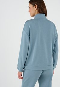 Felpa in pile azzurro chiaro con colletto alto, spalle scese e polsini a costine. Tessuto morbido e vestibilità casual.