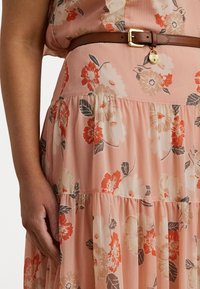 Blomstrete kjole i myk rosa stoff, med oransje og beige blomster, en lagdelt design, og et brunt belte med en gullspenne og charm.