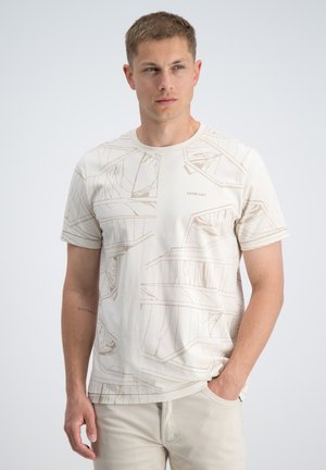 Man die een beige t-shirt met abstract patroon draagt en lichte broek, staat met één hand in de zak, kijkt zijwaarts tegen een effen achtergrond.