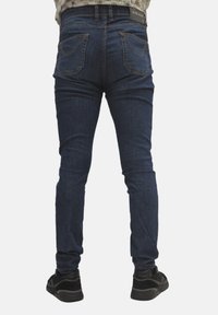 Jeans in denim blu scuro slim fit con vita media, cinque tasche e dettagli sul retro, abbinati a scarpe da ginnastica nere.