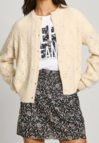 Cardigan de punto color crema con un patrón texturizado, con cuello redondo, cierre de botones y mangas holgadas, llevado sobre un vestido de estampado.