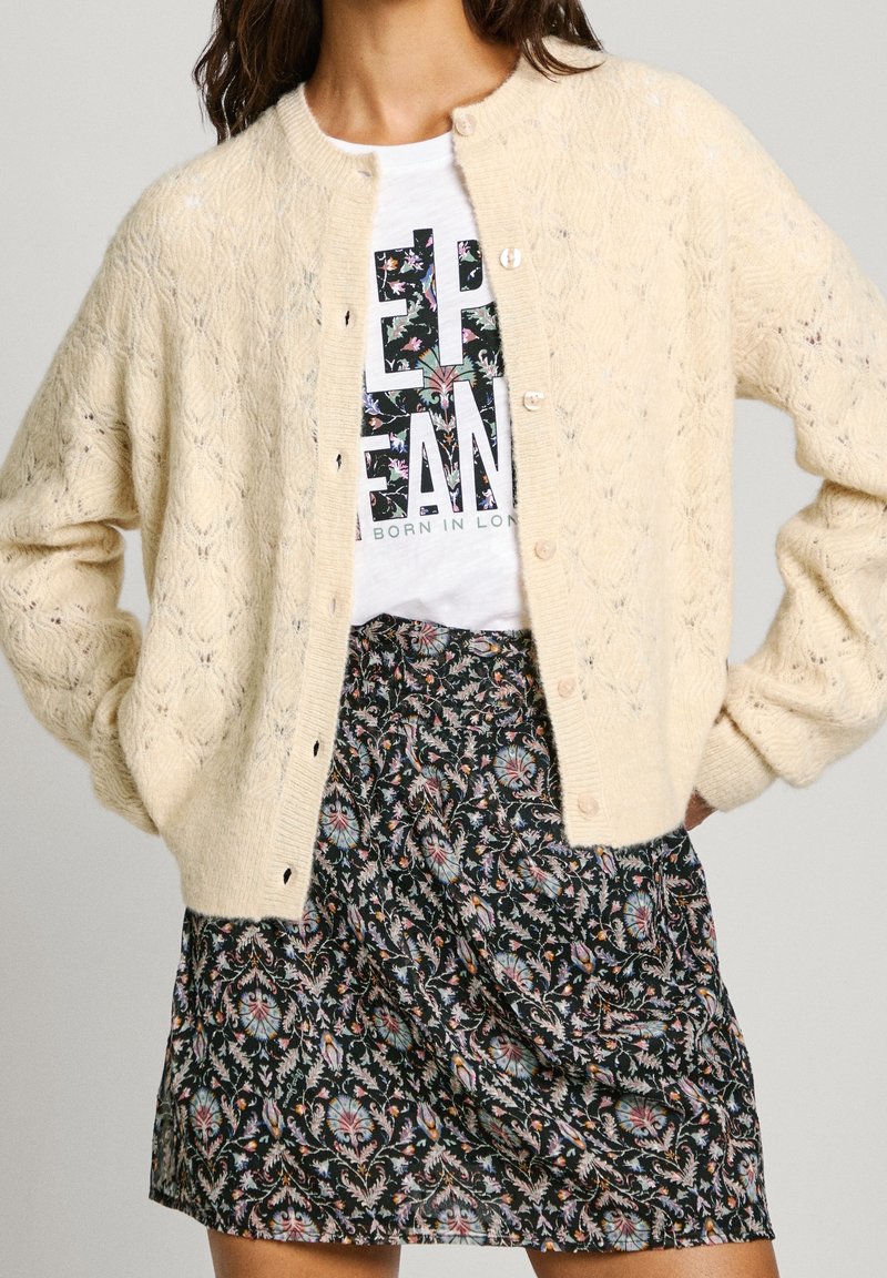 Cardigan de punto color crema con un patrón texturizado, con cuello redondo, cierre de botones y mangas holgadas, llevado sobre un vestido de estampado.