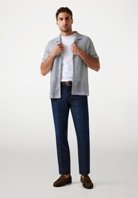 Camisa de manga corta, con botones, de rayas azules y blancas, llevada sobre una camiseta blanca lisa, combinada con jeans azul oscuro y mocasines marrones.