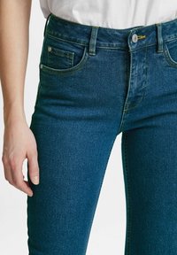 Džínové kalhoty s vysokým pasem v tmavě modré barvě s žlutým prošíváním. Mezi rysy patří pět kapes a přiléhavý střih s hladkým povrchem.