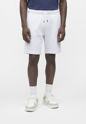 Homme portant un short blanc à cordon, des chaussettes blanches et des baskets blanches, debout devant un fond uni clair.